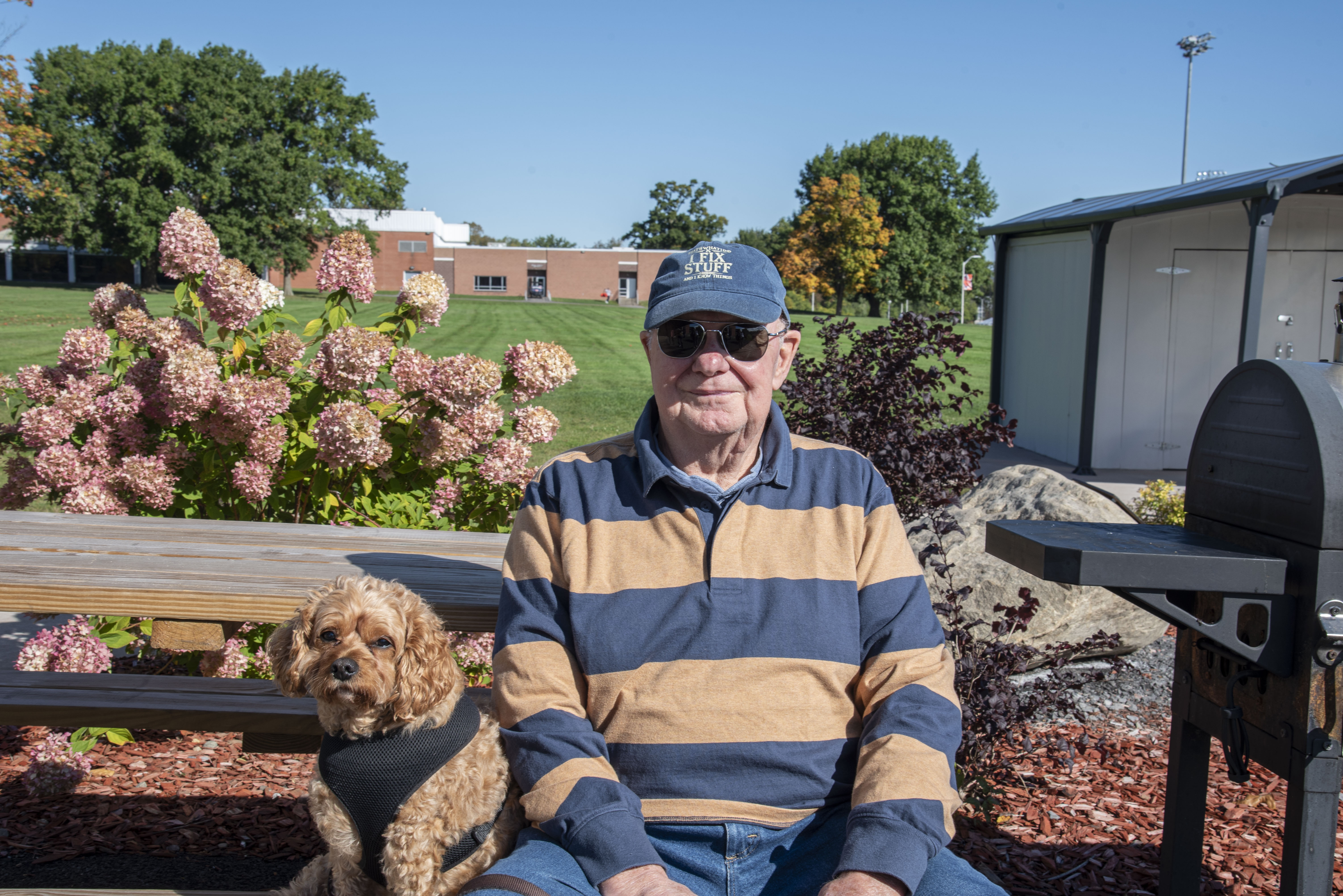 David Dubbelde in cap, orange and blue striped shirt, and sunglasses sits with his dog sit on a bench at Homecoming 2026 Construction Management BBQ.