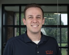 Tyler Davis in a blue Utica University polo shirt, stands in front of a window and smiles.