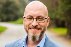 Joshua White, with beard and wearing glasses and a blue shirt and suit, smiles at the camera.