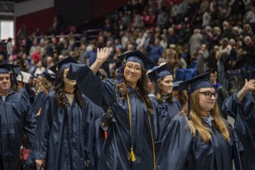 A student in a line of graduates in blue gowns and caps, waves.