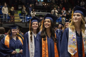 Row of students in blue commencement gowns and caps stand in a row in an auditorium.