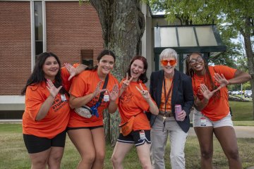 Students in orange t-shirts make moose ears with their hands, standing with President Stephanie Nesbitt in orange sunglasses.