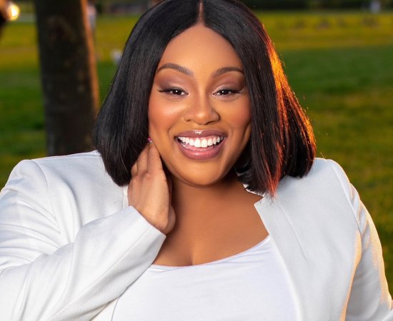 Akira Armstrong, in a white top and white blazer, stands outside on green grass, with her hand held up to her neck and smiles.