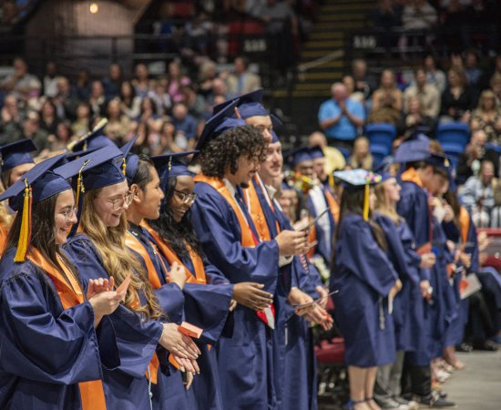 A row of graduates in blue commencement gowns and caps in an auditorium.