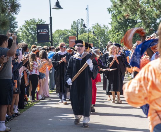 Professor Thomas Rossi acts as Macebearer, leading a procession of faculty down the sidewalk surrounded by cheering students.