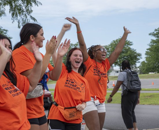 Students in orange t-shirts happily shout and wave their arms to greet newcomers to campus.
