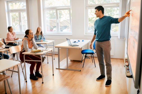 A teacher points to the board at the front of a classroom as students at desks listen.