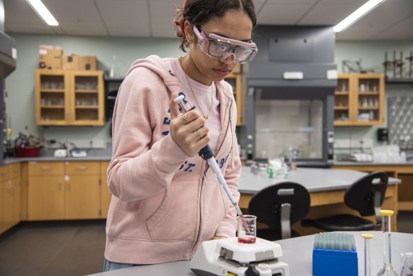Yamilet Taveras ’27, a chemistry major wearing a pink hoodie and safety goggles, pours a substance into a beaker in the chemistry lab.