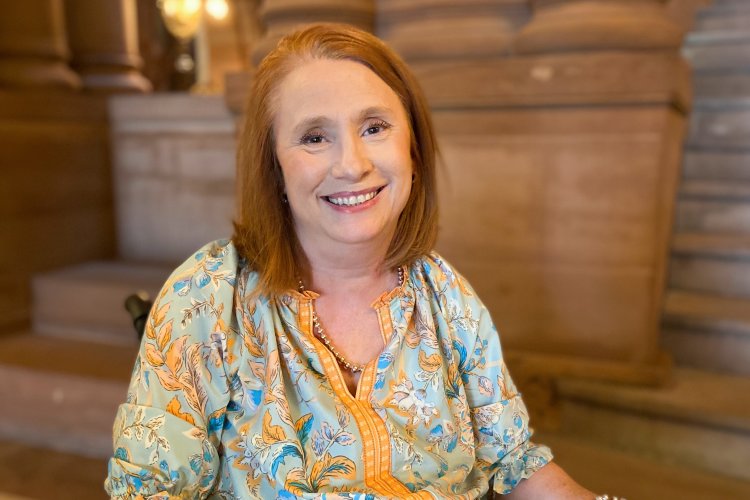 Kimberly Hill, with red hair and a blue-yellow shirt, sits in her wheelchair inside the capital building in Albany, smiling.