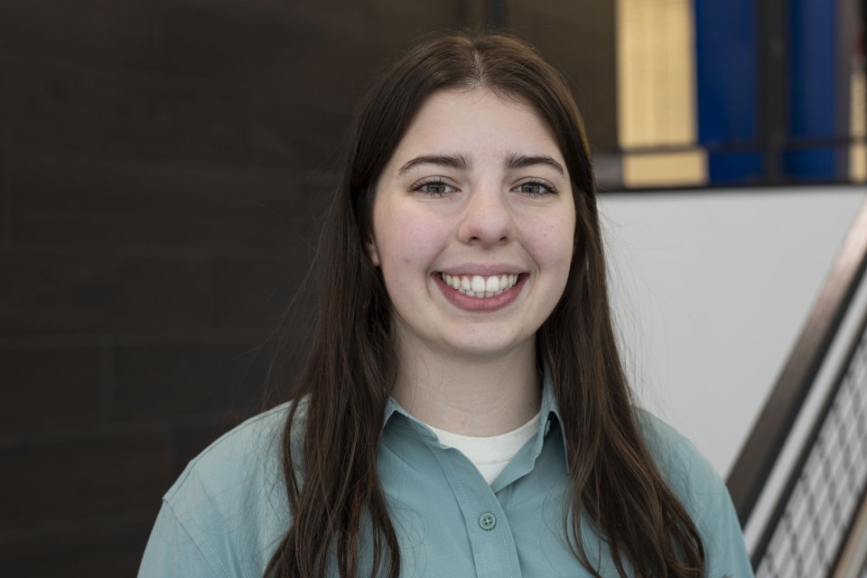 Sydney Kinsella, with long dark hair and wearing a blueish-green button-down top, looks at the camera and smiles.