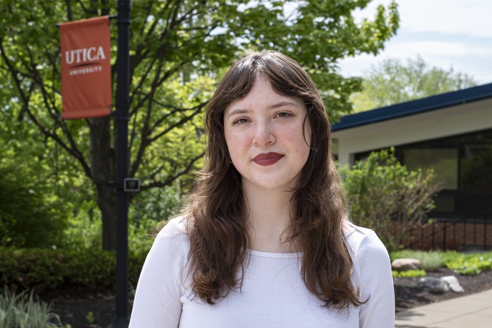 Bella Stephens, in white top with brown hair down past shoulders, stands in front of foliage on campus.